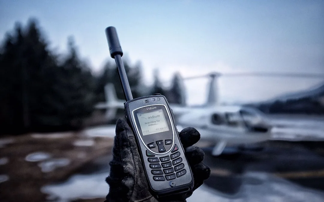 Gloved hand holding an Iridium satellite phone outdoors in winter, with a small plane behind